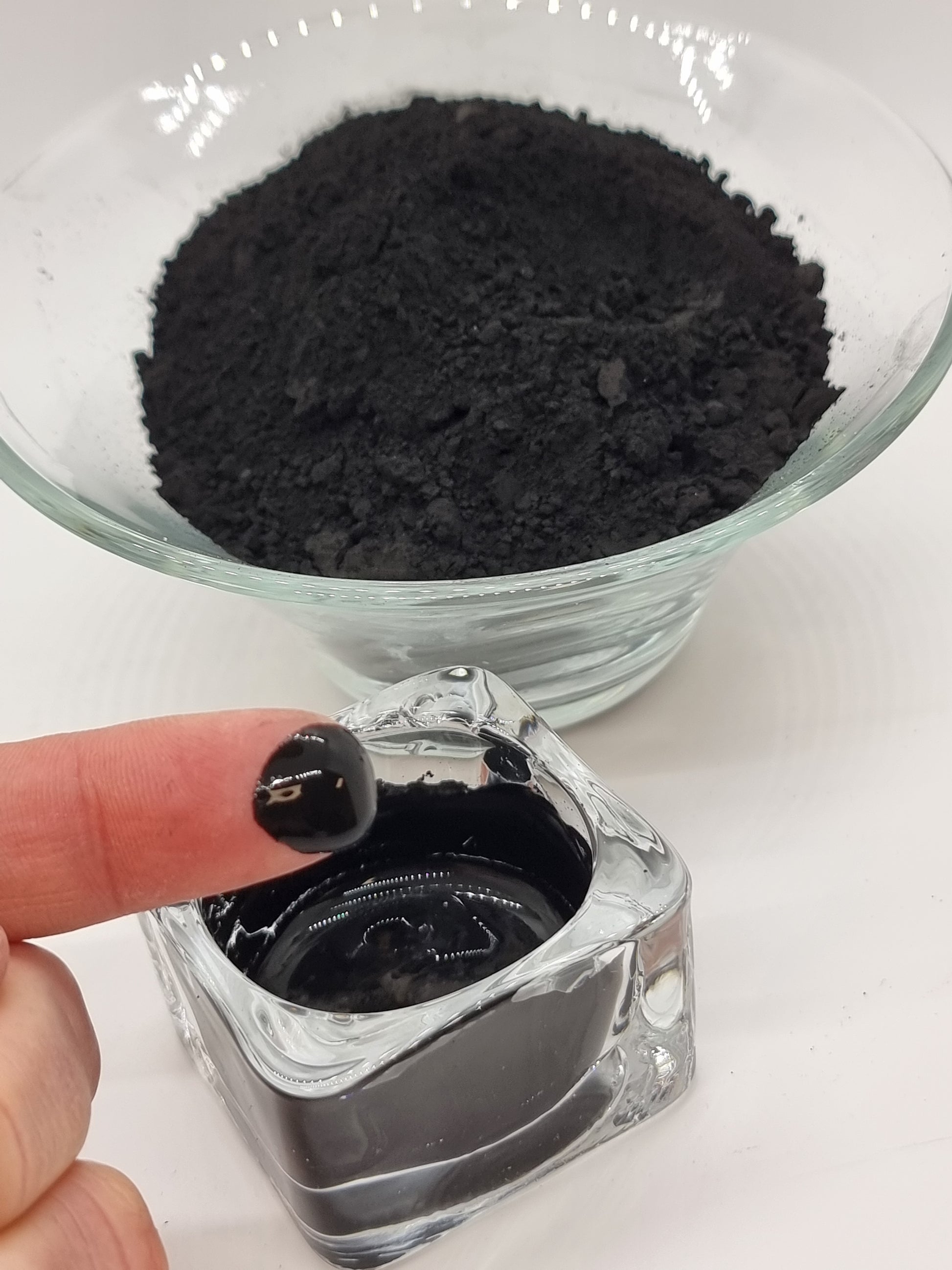 glass bowls one with dry black powder, activated charcoal, and the other with charcoal paste. A hand can be seen with a finger that has a blob of charcoal paste on it that catches the light and appears shiny