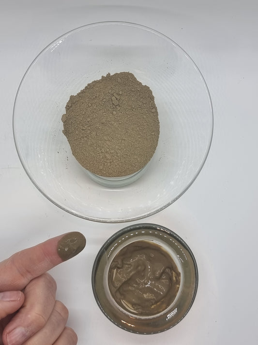 two glass bowls, one with fine brown grey dead sea mud powder that is dry and one with dead sea mud powder that has been mixed with water to a shiny looking paste. A hand is showing in the corner with a finger that has been dipped into the wet dead sea mud