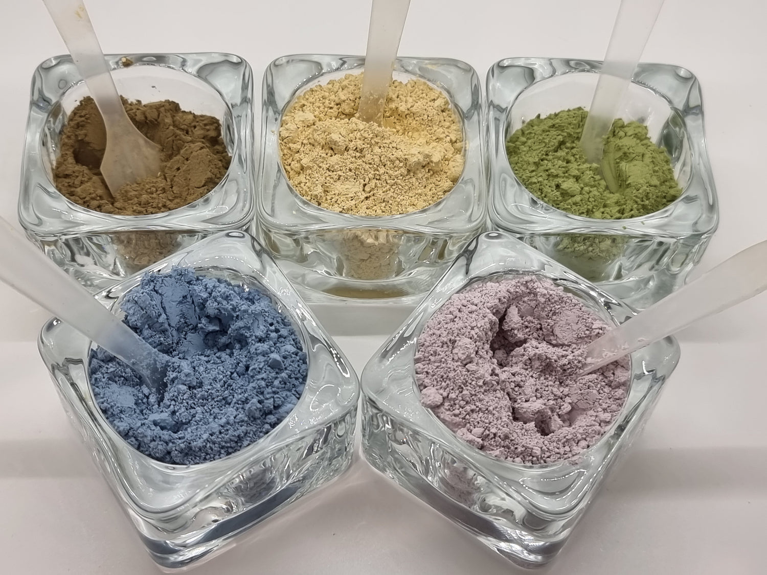 Cosmetic grade clays in glass pots with spoons featuring blue, pink, yellow, green and brown naturally coloured powders