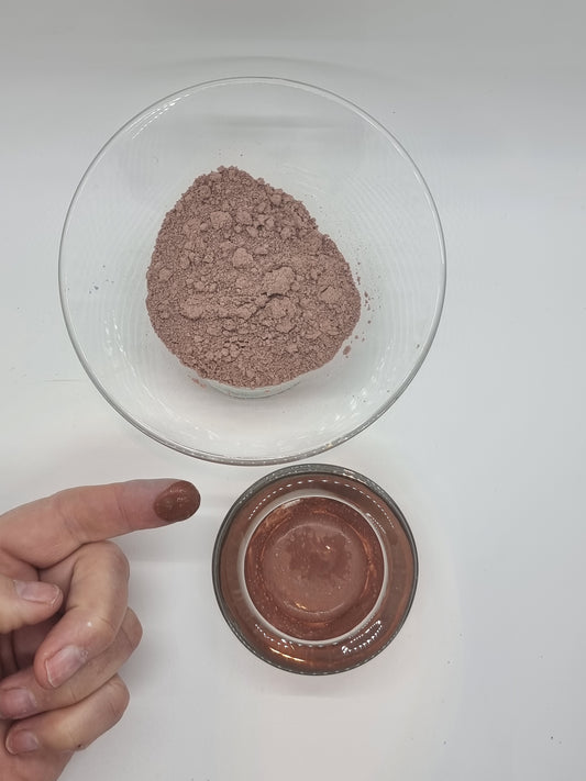 two glass bowls one containing fine french pink clay dry powder, the other pink clay paste. A hand is visible with a finger that has a blob of wet pink clay on the end.