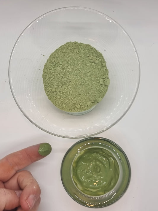 two glass bowls, the larger one containing dry glacial marine clay that is a mid green colour, the smaller holding clay that has been mixed to a paste with water, and reflects the light. A hand can be seen with a finger outstretched that has been dipped into the wet clay, visible on the end of the finger as a shiny green paste