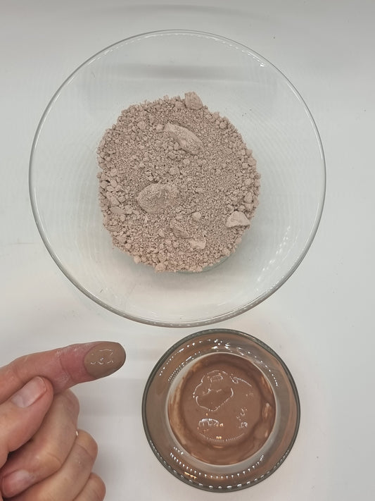 Moroccan Nude clay in two glass bowls. Light pink powder in one large bowl and darker pink wet clay paste in the other. A hand is visible with a finger that has a blob of wet clay on it. Light shines on the wet clay.