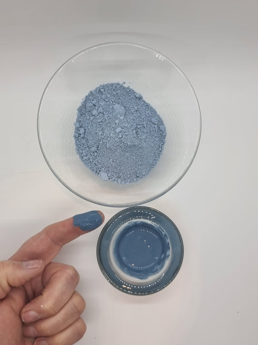 Serbian Blue clay in powder form and mixed with water showing the bright blue colour of the clay. A hand is visible with a finger pointing that has a blob of wet blue clay on it. Light reflects on the wet clay.